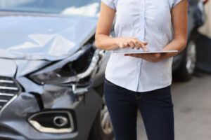 A woman standing in front of a damaged car, using a tablet to document the accident.