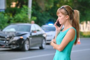 A woman standing on the street, speaking on the phone after a car accident with a damaged black car in the background.