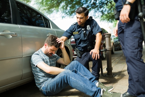 A police officer assisting an injured man sitting on the ground beside a parked car after an accident, with the officer offering support.