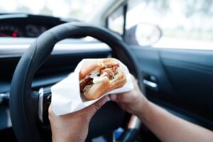 A person eating a sandwich while driving, one hand on the wheel and the other holding food, representing distracted driving due to eating.